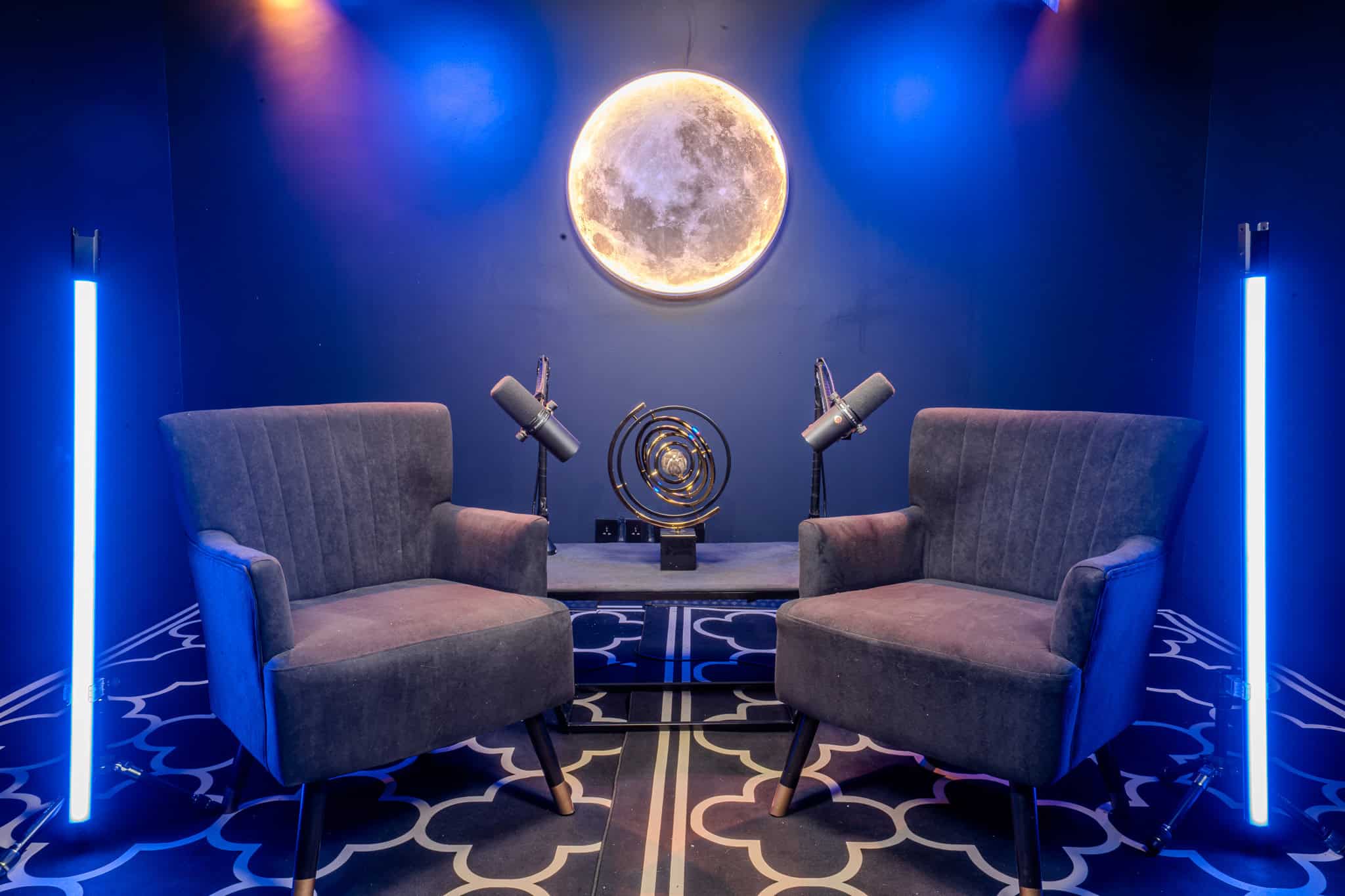 Brightly lit podcast studio featuring moon wall decor, two comfortable chairs, and professional microphones.